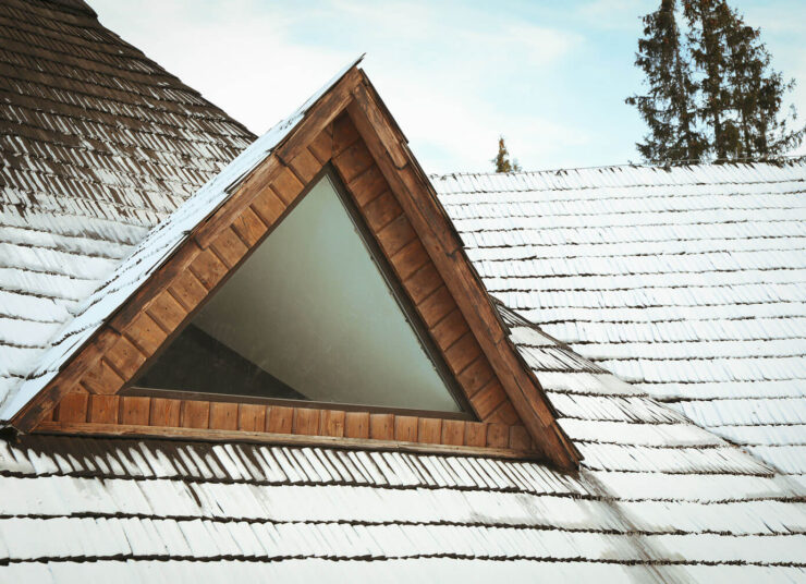 Eine dreieckige hölzerne Dachgaube mit Glas ist in ein leicht mit Schnee bestäubtes Schindeldach eingelassen. Im Hintergrund sind immergrüne Bäume unter einem teilweise bewölkten Himmel zu sehen.