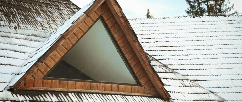Eine dreieckige hölzerne Dachgaube mit Glas ist in ein leicht mit Schnee bestäubtes Schindeldach eingelassen. Im Hintergrund sind immergrüne Bäume unter einem teilweise bewölkten Himmel zu sehen.
