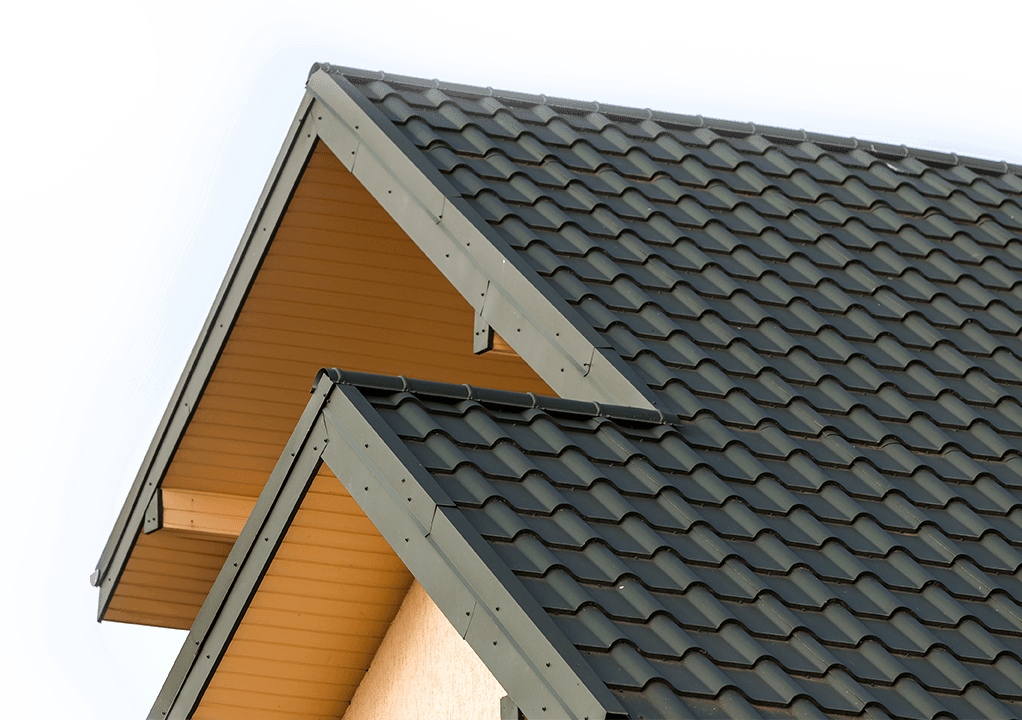 Nahaufnahme eines Hausdachs mit dunkelgrauen Metalldachziegeln und zwei Giebelspitzen. Die Traufe und die Unterseite des Daches sind mit hellen Holzplatten verkleidet. Der Himmel ist bewölkt und weiß.