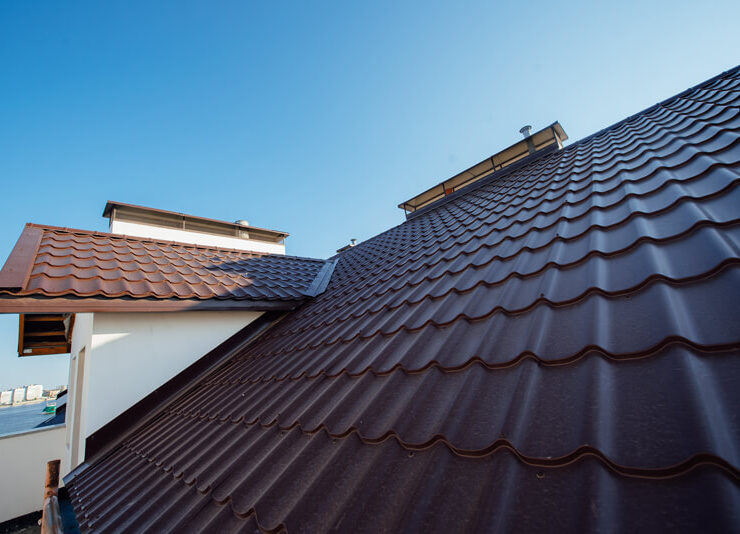 Nahaufnahme eines mit braunen Metalldachziegeln gedeckten Hausdachs bei klarem blauem Himmel. Die Dachplatten haben ein wellenförmiges, ineinander greifendes Muster. Ein Teil eines anderen Dachabschnitts und eine weiße Wand sind ebenfalls zu sehen.