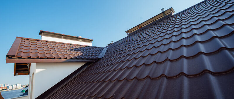 Nahaufnahme eines mit braunen Metalldachziegeln gedeckten Hausdachs bei klarem blauem Himmel. Die Dachplatten haben ein wellenförmiges, ineinander greifendes Muster. Ein Teil eines anderen Dachabschnitts und eine weiße Wand sind ebenfalls zu sehen.