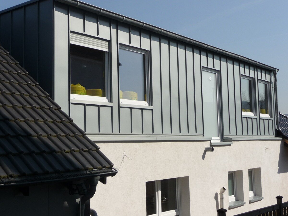 Dachdecker Ein modernes Haus mit hellgrauer vertikaler Metallverkleidung im Obergeschoss und weißem Stuck im Erdgeschoss, mit großen rechteckigen Fenstern und einem schrägen dunklen Dach im Vordergrund.