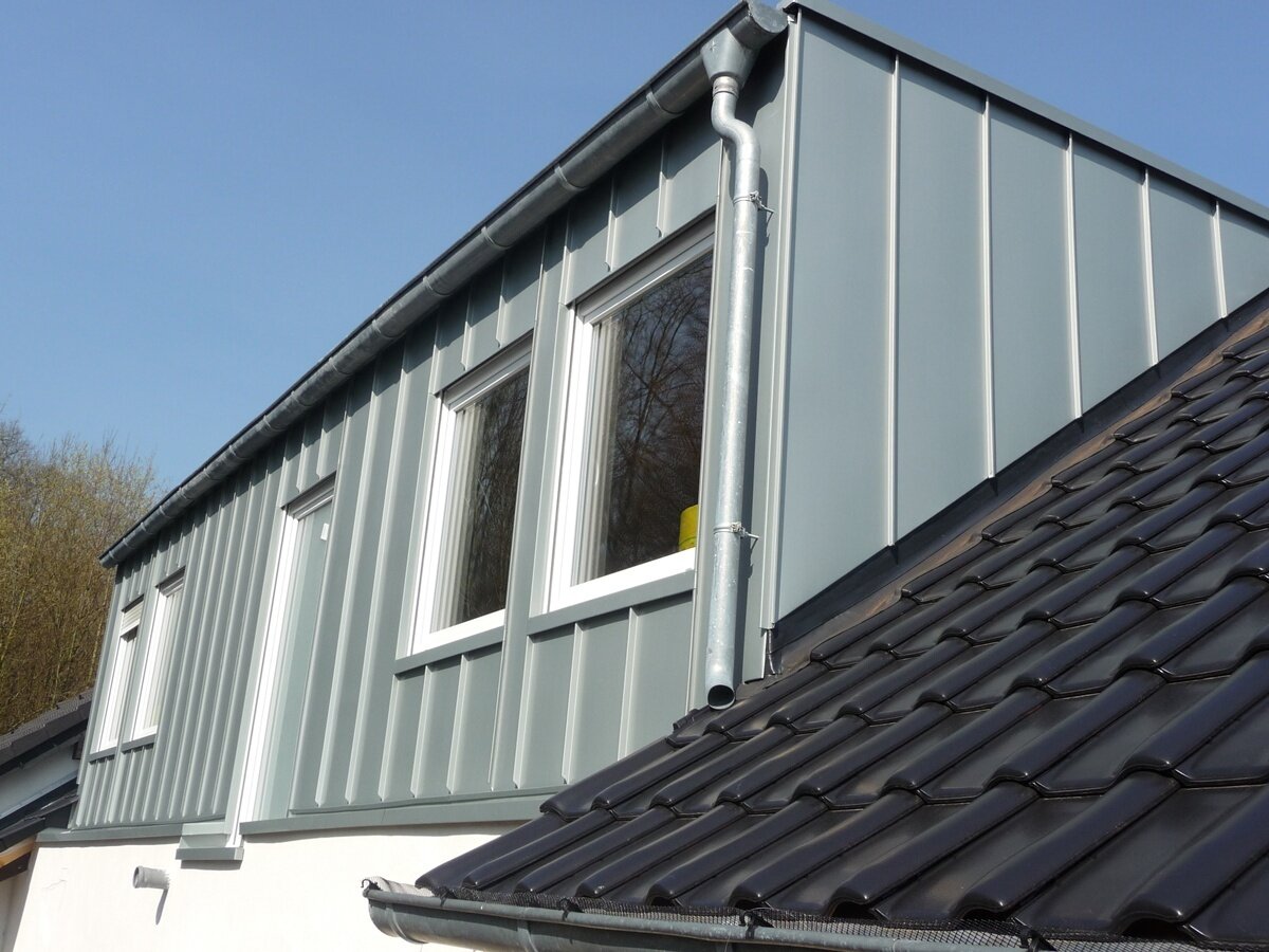 Dachdecker Eine Nahaufnahme eines modernen Hauses mit vertikalen grauen Metallverkleidungen, zwei weiß gerahmten Fenstern, einer Regenrinne aus Metall und einem dunkelgrauen Ziegeldach unter einem klaren blauen Himmel.