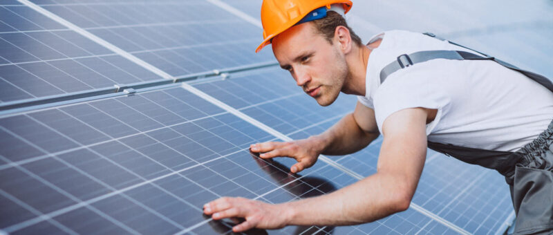 Ein Arbeiter, der einen Schutzhelm und einen Overall trägt, inspiziert oder wartet Solarmodule, wobei er sich über die Oberfläche beugt und diese berührt, so dass Reihen von Solarmodulen sichtbar sind.