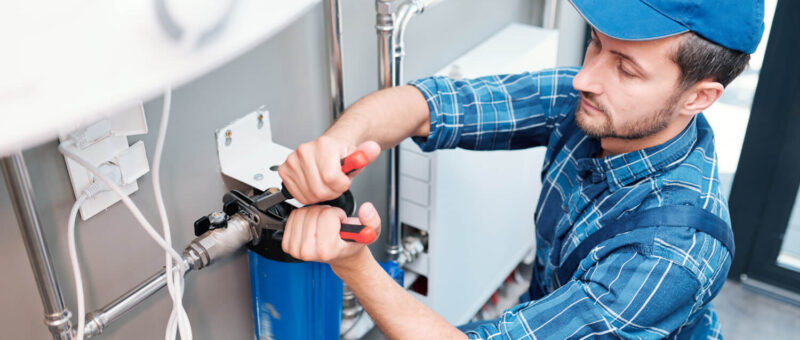 Ein Klempner mit blauer Mütze und kariertem Hemd benutzt einen Schraubenschlüssel, um ein Wasserfiltersystem an einer Wand mit sichtbaren Rohren zu installieren oder zu reparieren.