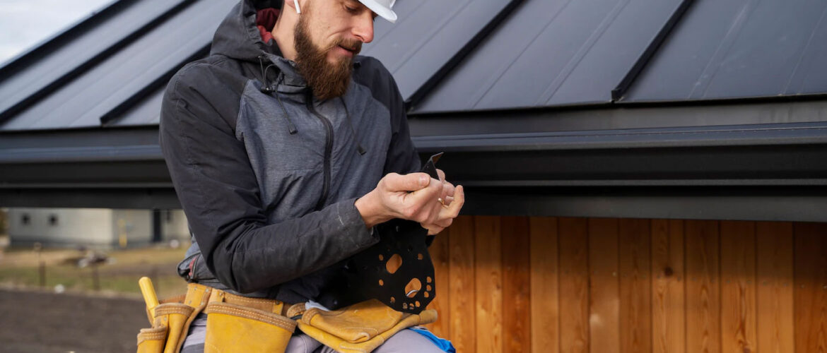 Ein Bauarbeiter mit weißem Schutzhelm und Werkzeuggürtel kniet an einem Holzgebäude mit Metalldach und prüft oder bereitet ein kleines Werkzeug oder Befestigungsmittel in seinen Händen vor.