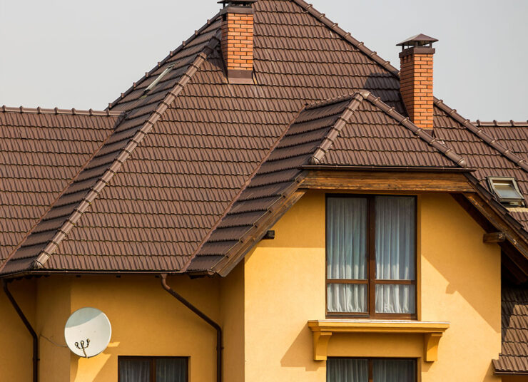 Ein modernes Haus mit gelben Wänden, großen Fenstern mit Vorhängen, braunen Dachziegeln, zwei gemauerten Schornsteinen und einer weißen Satellitenschüssel an der Wand.