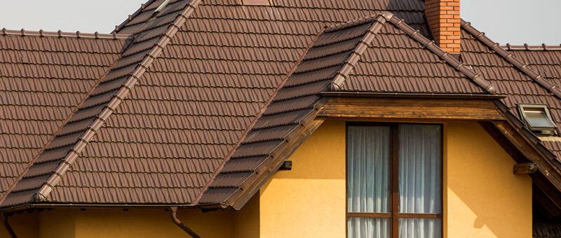 Ein modernes Haus mit gelben Wänden, großen Fenstern mit Vorhängen, braunen Dachziegeln, zwei gemauerten Schornsteinen und einer weißen Satellitenschüssel an der Wand.