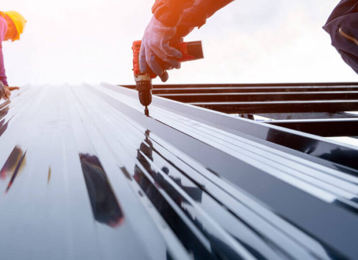 Nahaufnahme eines Arbeiters, der mit einer Bohrmaschine glänzende Metalldachplatten auf einem Gebäude anbringt. Im Hintergrund ist ein weiterer Arbeiter zu sehen, beide tragen Schutzkleidung.