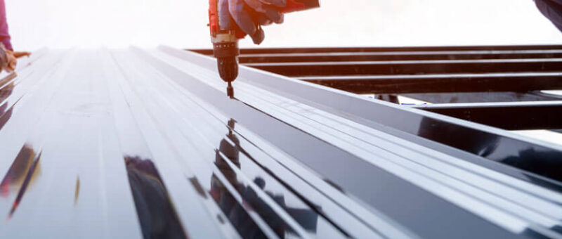 Nahaufnahme eines Arbeiters, der mit einer Bohrmaschine glänzende Metalldachplatten auf einem Gebäude anbringt. Im Hintergrund ist ein weiterer Arbeiter zu sehen, beide tragen Schutzkleidung.