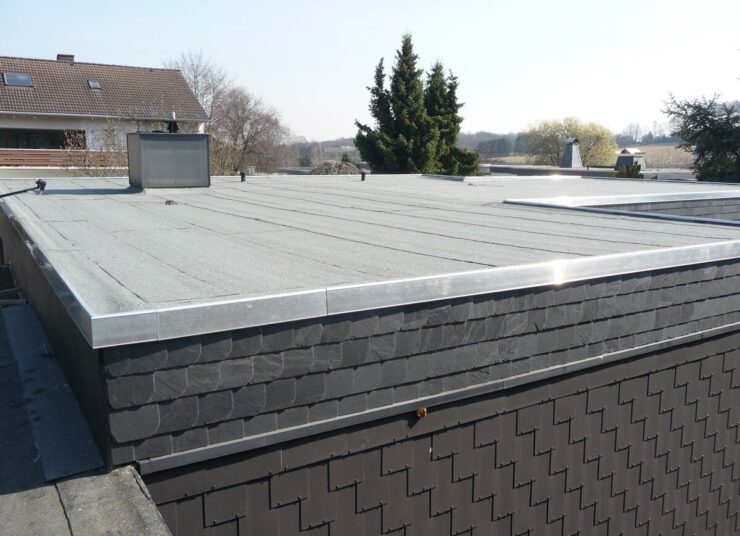 Es ist ein Flachdach mit einer hellen, glatten Oberfläche und einer Metallumrandung zu sehen. Das Gebäude hat eine dunkle, mit Schindeln gemusterte Verkleidung. Im Hintergrund sind bei klarem Himmel Bäume und benachbarte Häuser zu sehen.