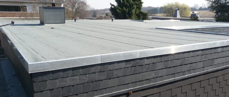 Es ist ein Flachdach mit einer hellen, glatten Oberfläche und einer Metallumrandung zu sehen. Das Gebäude hat eine dunkle, mit Schindeln gemusterte Verkleidung. Im Hintergrund sind bei klarem Himmel Bäume und benachbarte Häuser zu sehen.